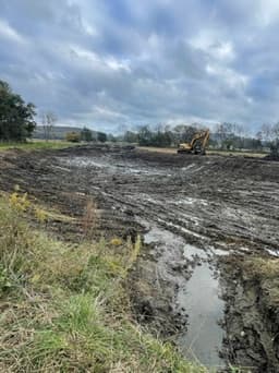 Wide pond excavation in progress with excavator visible and standing water